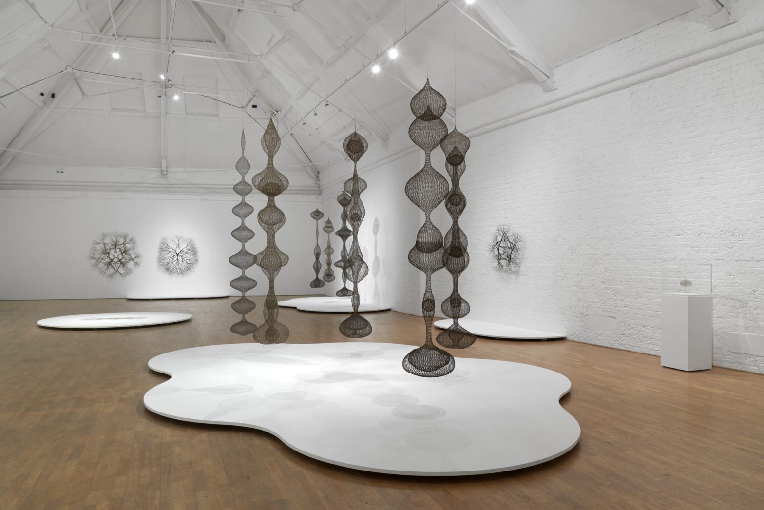 Several sculptures hanging from a high ceiling. They are made of looped wire and form long vertical abstract bulbous shapes. A white flat abstract form is on the floor beneath them. Wire sculptures hang on the walls behind them.