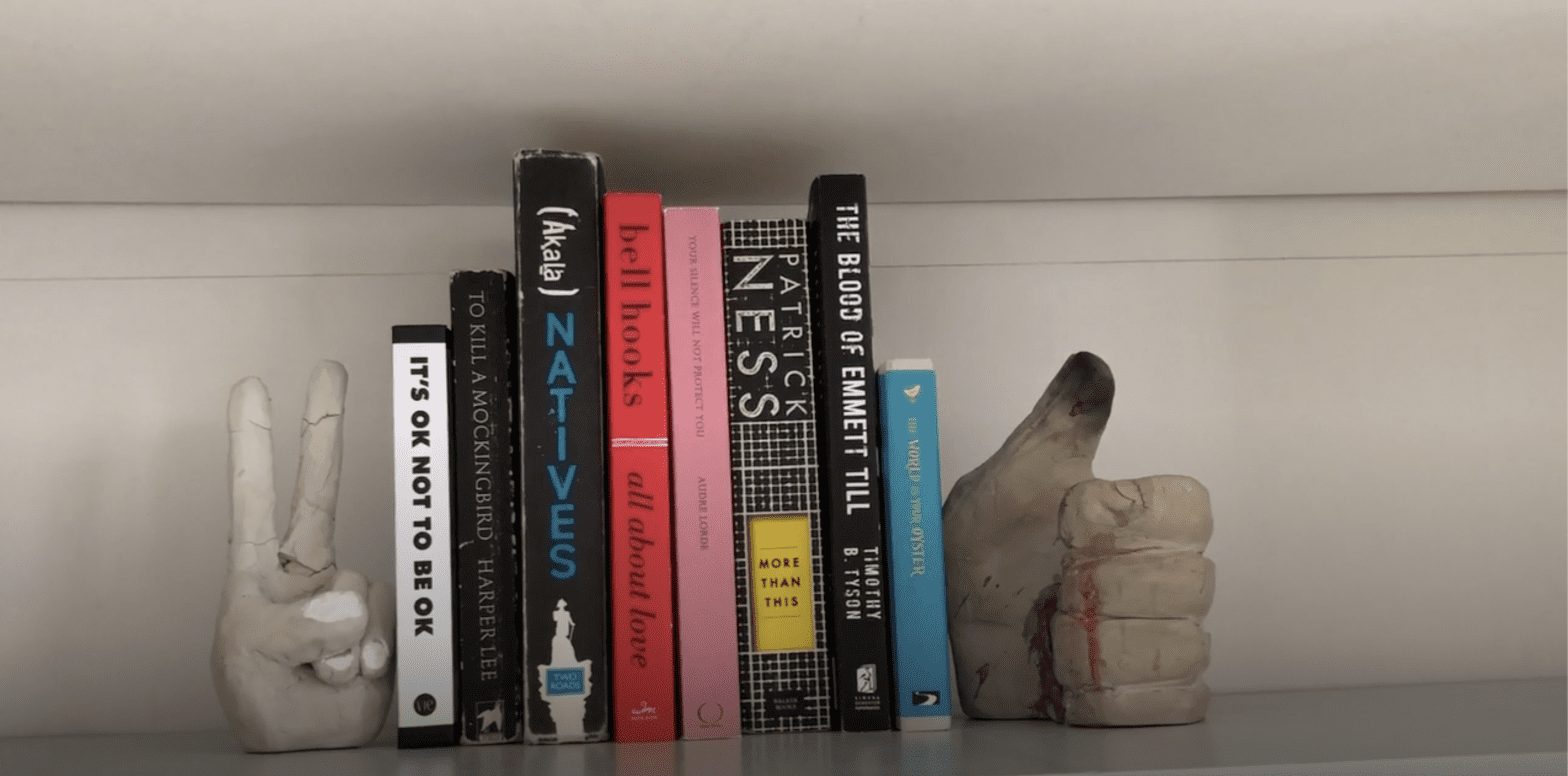 A bookshelf with several books displayed: It's OK Not to be OK; To Kill a Mockingbird by Harper Lee; Natives by Akala; All About Love by bell hooks; Your Silence Will Not Protect You by Audre Lorde; More Than This by Patrick Ness; The Blood of Emmett Till by Tiimothy B Tyson; and The World is Your Oyster. The books are framed by two book ends, one in the shape of a thumbs up, and one with the fingers doing a peace sign