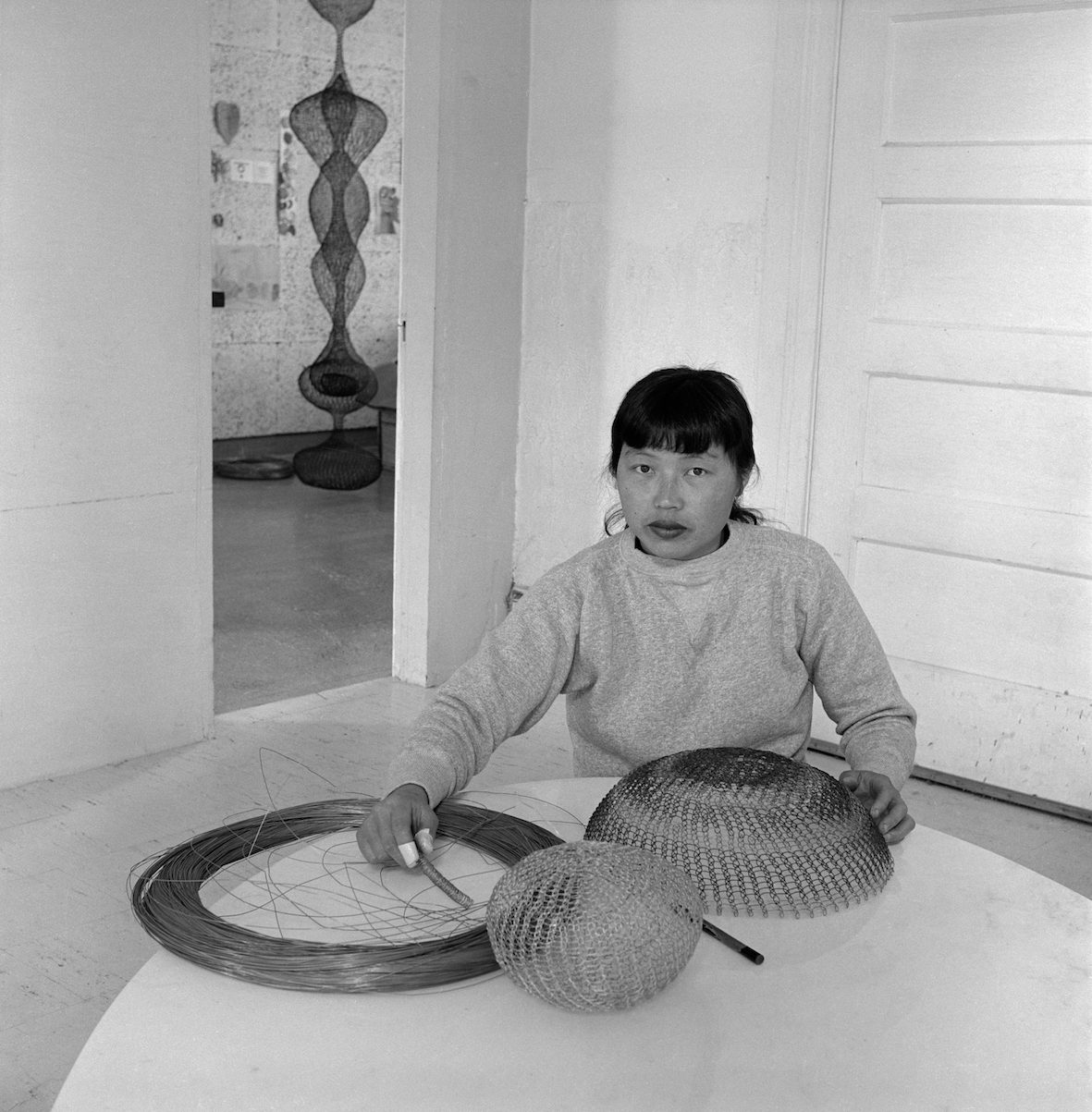 A young woman creating circular wire sculptures from large loops of wire. Asawa is sitting and stares. The background features a hanging looped wire sculpture in another room.