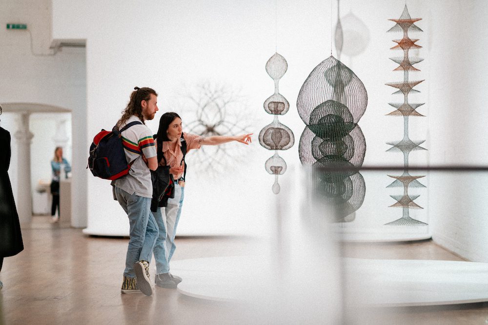 Two people viewing a series of hanging sculptures made of looped wire, in organic round shapes and layered disk shapes that overlap. One of the people is pointing to the sculpture.