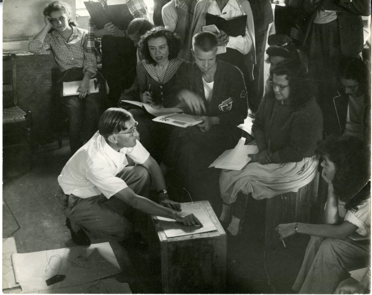Black and white photo of a classroom, with young people sitting around on chairs and boxes watching the teacher (Josef Albers) who is crouching down at the front demonstrating something on a piece of paper.