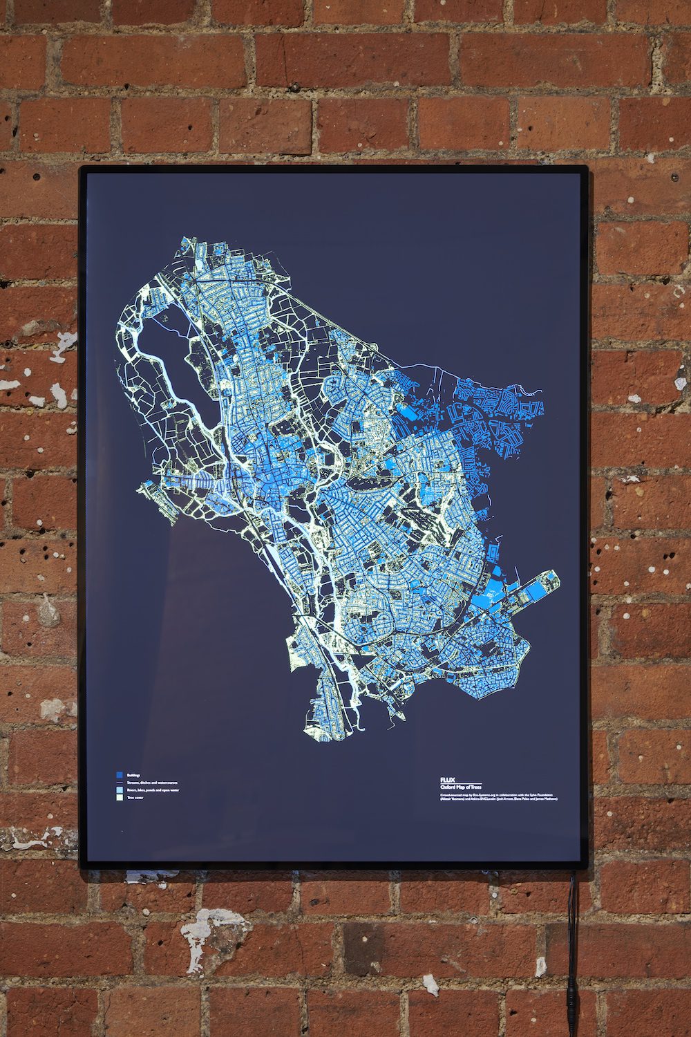 Framed map on a brick wall. It is an aerial map of Oxford is in shades of blue and green.