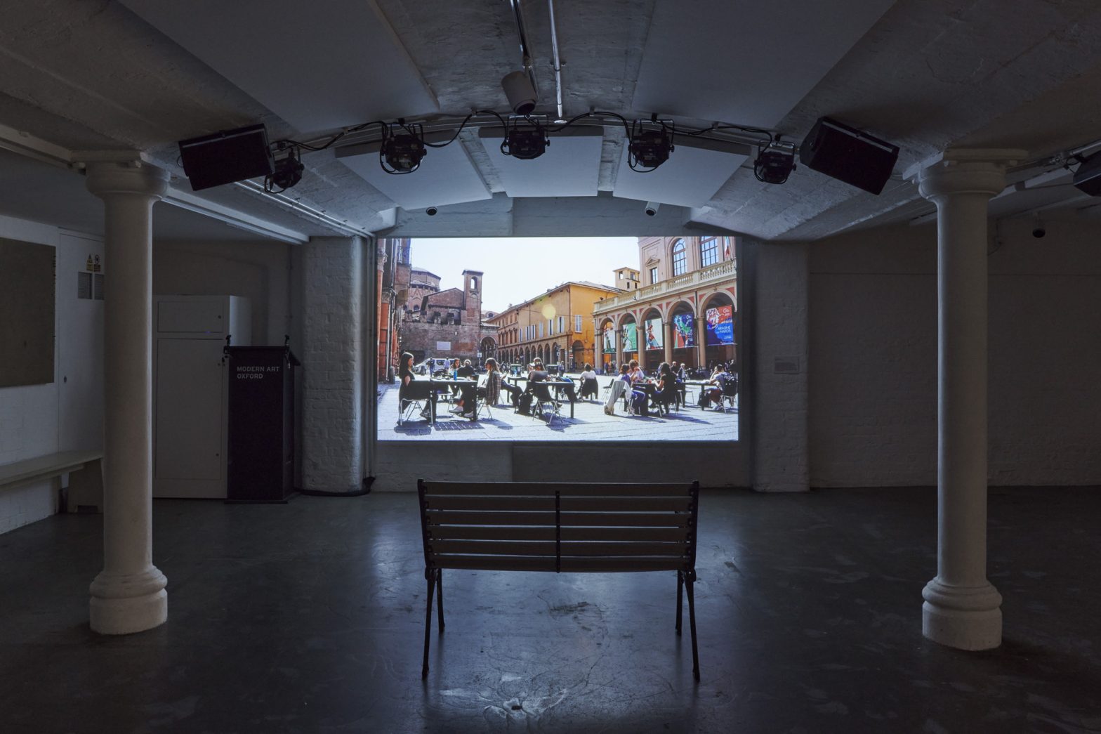 A screen with an image of a town square projected on it in a darkened room, framed by two white columns. A park bench is placed in front of the screen.