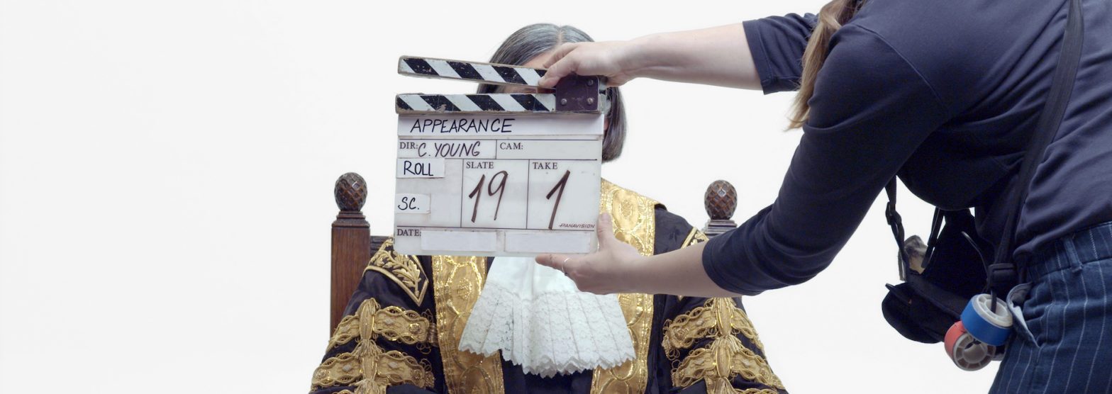 A film clapperboard being held in front of a person in an ornate gold and black costume, who is sitting with their hands in front of them against a white background.