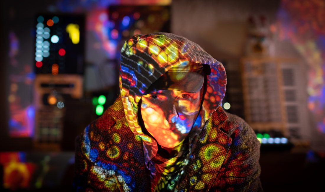 A man crouched down wearing a hoodie and jacket looking directly into the camera with very brightly coloured patches of light covering his body, with reflections of electronic equipment behind.