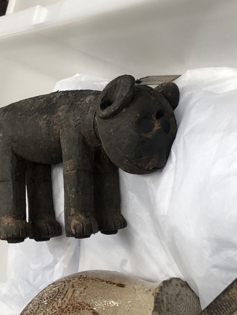 Old small stone carved animal artefact places on protective white material, with large round eye holes, oval ears and four straight legs with paws.
