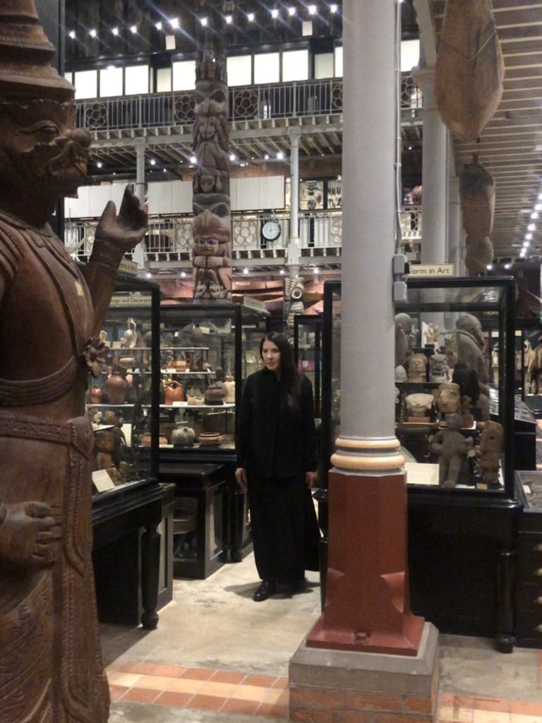Woman in a long black dress with black hair swept to one side in a museum filled with objects in cases, with a large stone sculpture on the left and a column going from floor to ceiling.