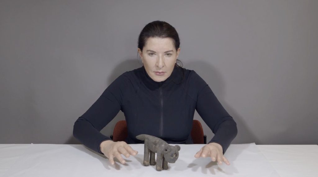 Older woman with black hair pulled back sitting at the table, with hands held out either side of a stone carving of an animal.