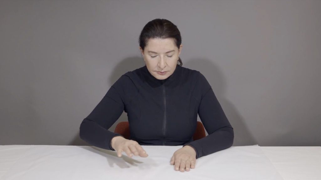 Older woman with black hair pulled back sitting at a white table with her eyes closed, with right hand resting palm down and the other hovering over the table.