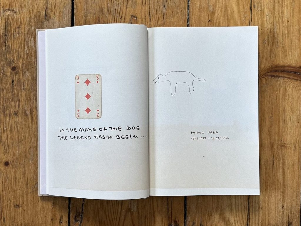 Open book with illustrations on a wooden floor, with a page featuring the three of diamonds playing card and a drawing of a dog, with the words: In the name of the dog the legend has to begin, my dog Alba 15.05.1977 - 18.12.1992