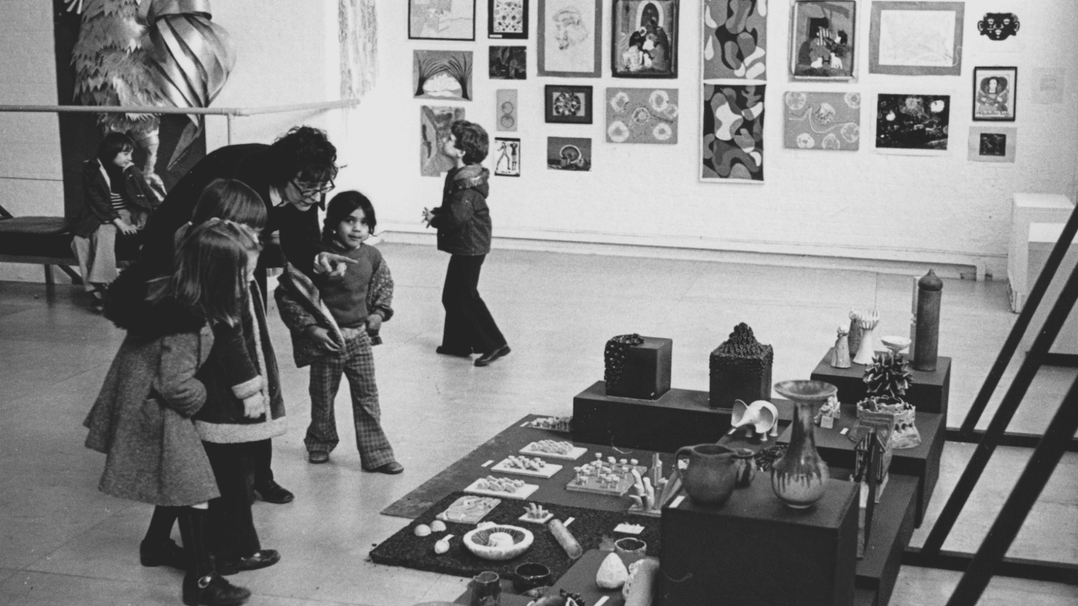 Black and white photo of an adult bending down to talk to three small children in an art gallery. The adult is pointing at a display of ceramic pots and objects laid out on the floor in front of them. In the background artworks are displayed on a white wall.