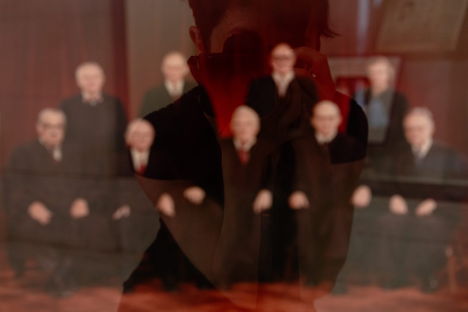 Group of formally dressed judges, featuring eight men and one woman against a red background. The image is overlaid with a reflection of the artist Carey Young taking a photograph.