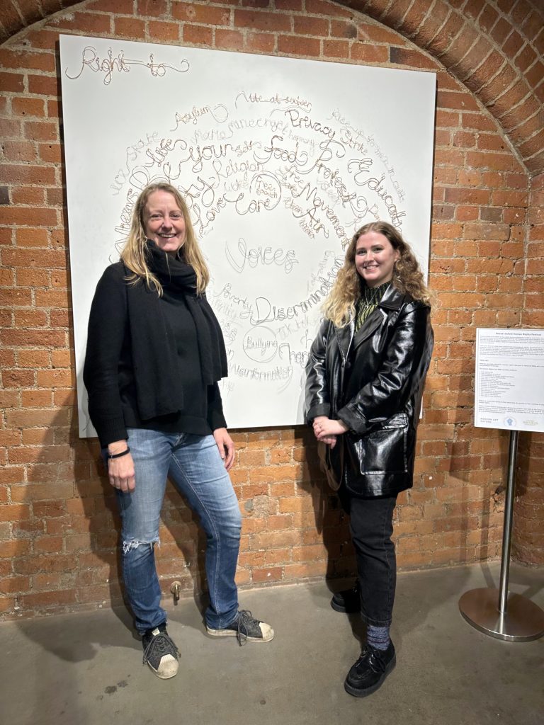 Two people standing in front of an artwork on a brick wall. They have long hair and are smiling. The artwork is a spiral of words made with wire on a white canvas. 