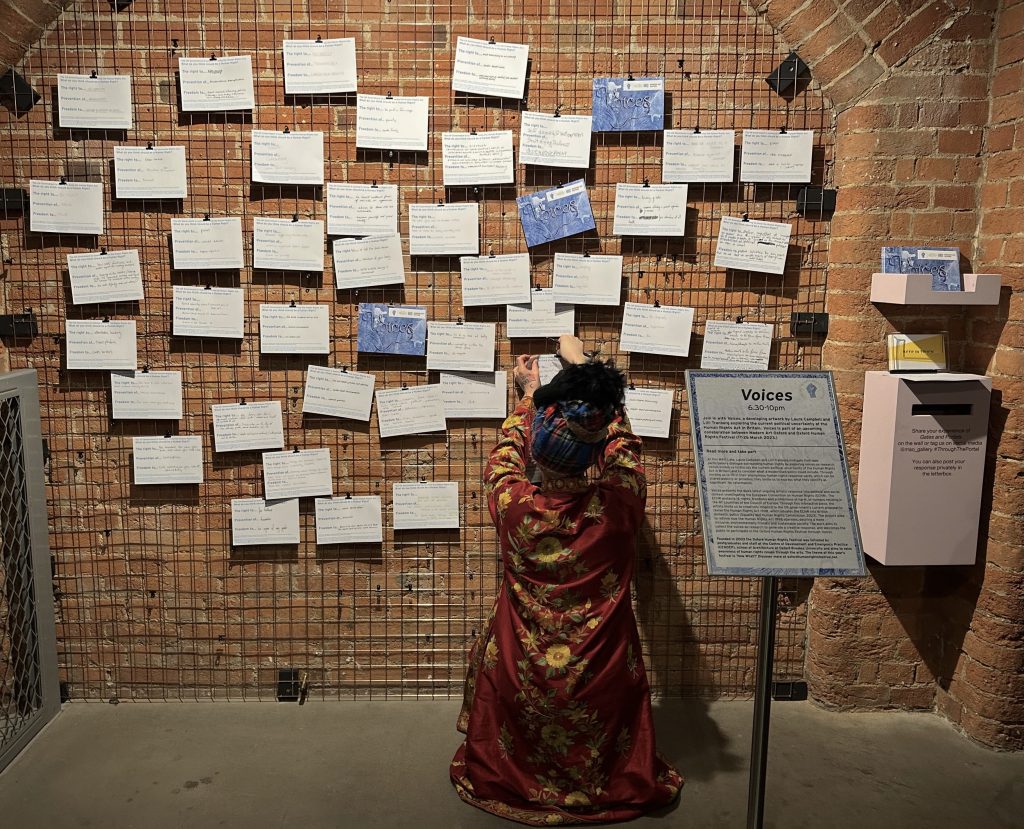 Wall of mesh wire with an arrangement of handwritten cards. A person in ornate clothing is hanging another card on the wall.
