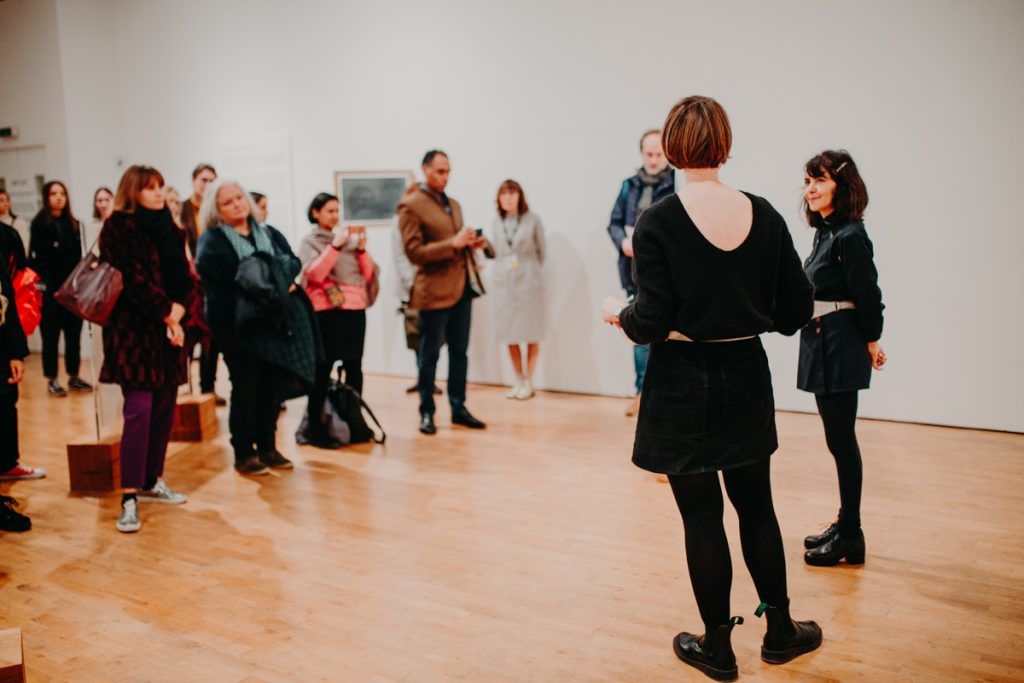 Group of people standing in Modern Art Oxford Upper Gallery. They are facing a woman with her back to the camera. They are listening to her talk.