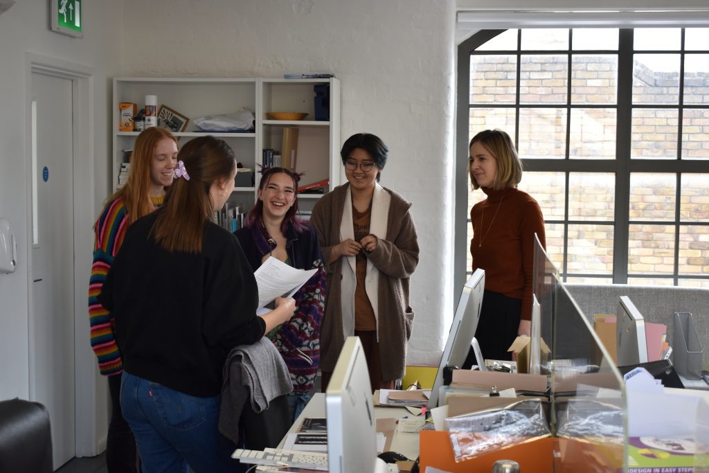 Four people stand together smiling in a bright office space with a large window, a woman in a black jumper holds a document.