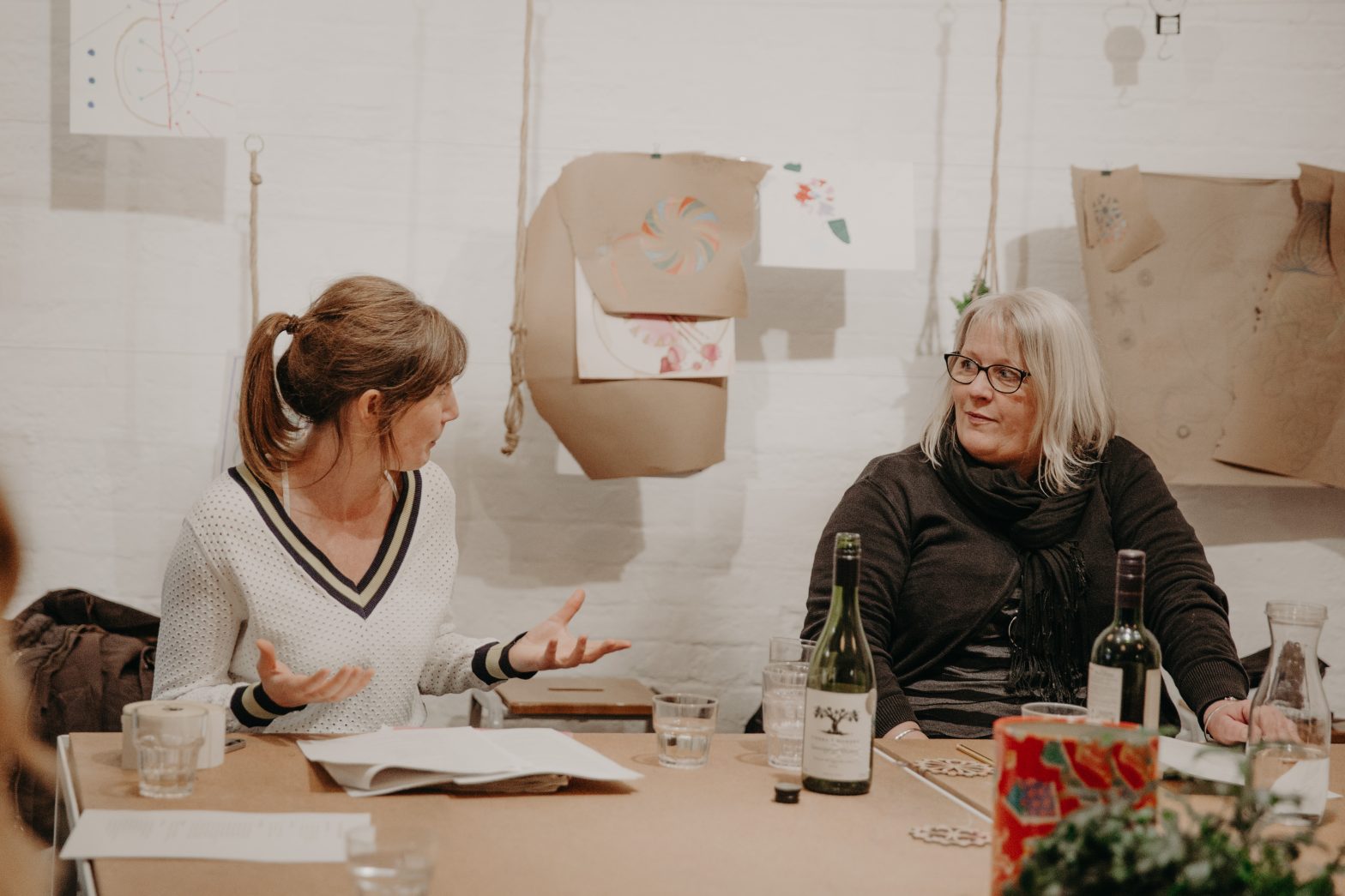 Two women engaged in a discussion in Modern Art Oxford's creative space. The woman on the left has a brown ponytail and is wearing a white jumper, she is gesturing with her hands. The other woman has blonde hair, wears glasses and a black jumper. There is art hanging up on the white walls behind them.