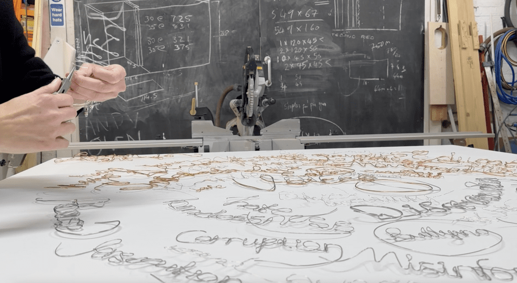 The hands of a person working on an artwork in a workshop. The artwork displays a spiral of words made with metal wire. The person is using small pliers to bend the wire.  There is a blackboard with numbers and calculations in the background. 
