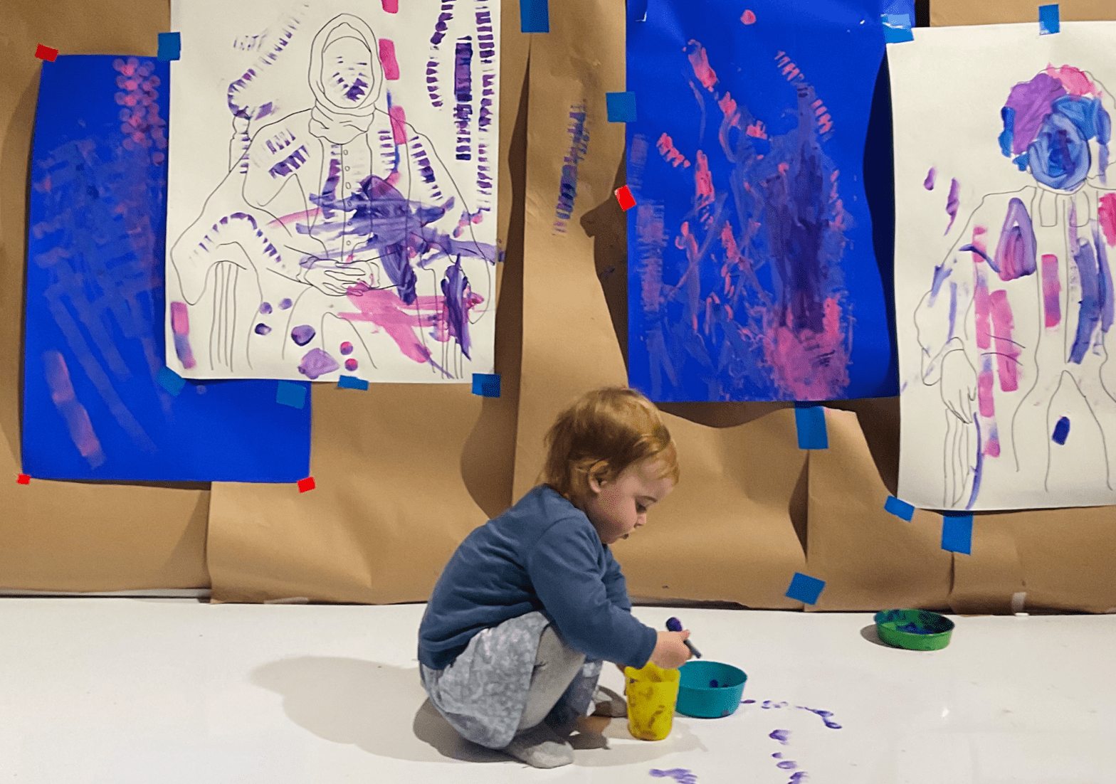 Small child painting on a floor covered in white paper. In the background paper hangs from the wall covered in pink and purple splatters of paint.