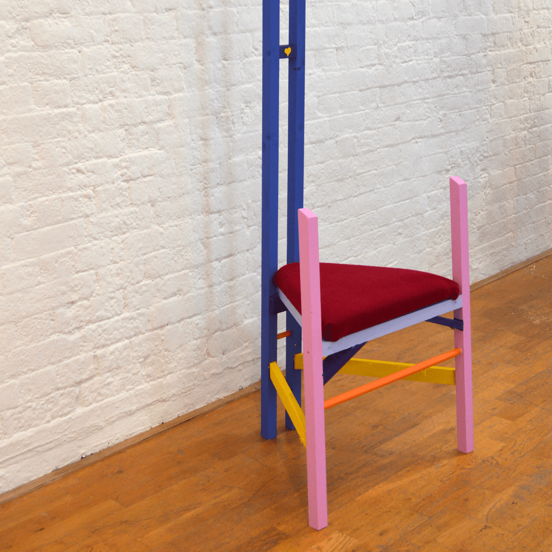 A tall wooden chair painted bright pink, yellow and blue. On the seat is a red velvet triangular cushion.
