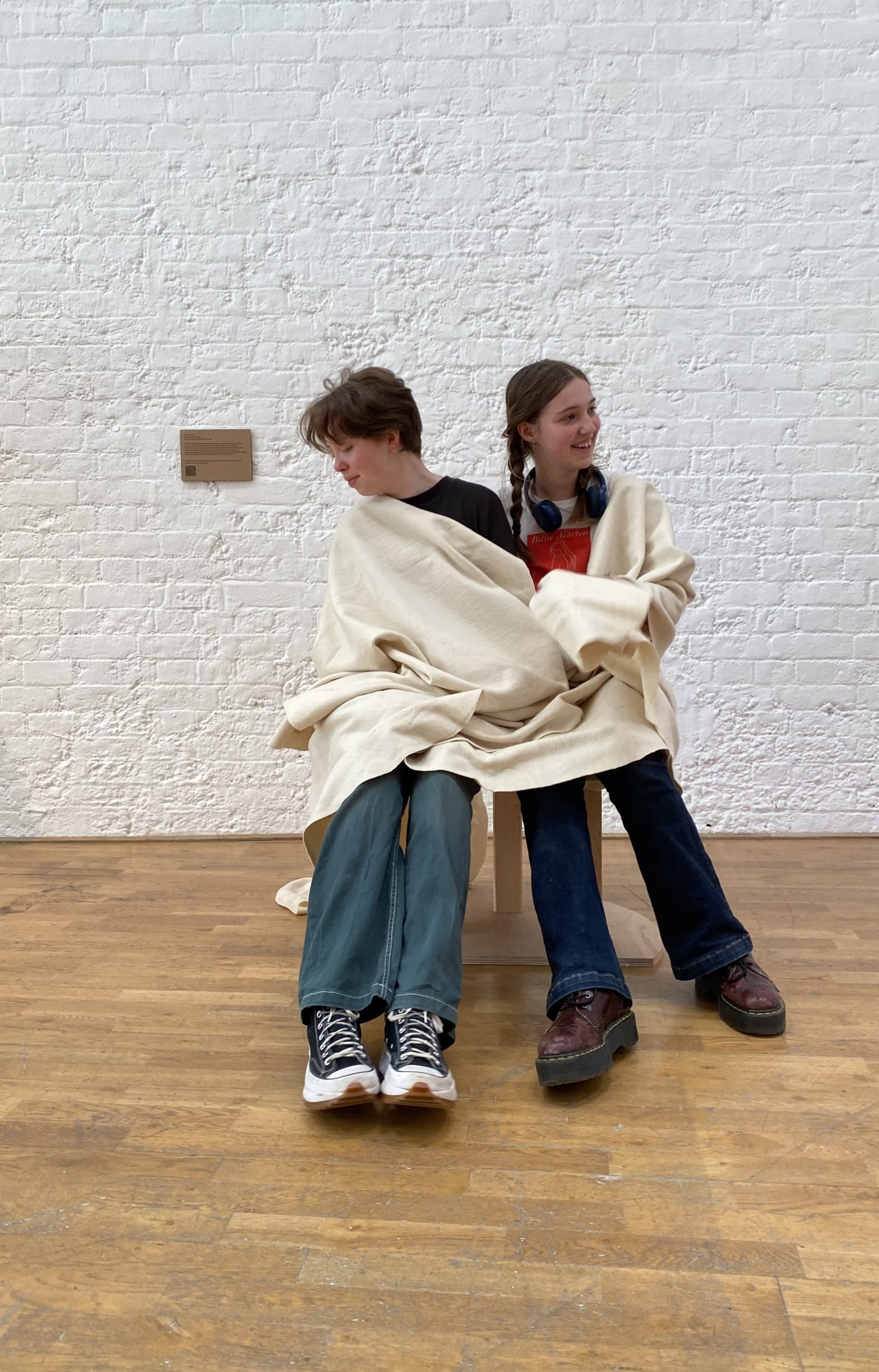 Two young people sitting on a chair that has a built in blanket. They have wrapped the blanket around themselves.