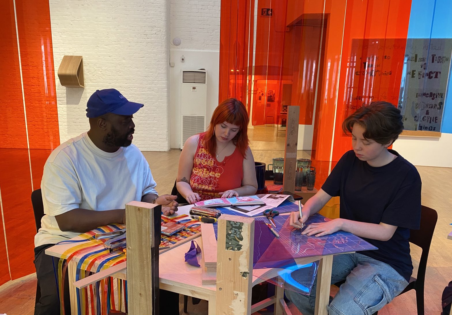 Three people sitting and working together at a table. Behind them coloured sheets of plastic hang down from the ceiling.