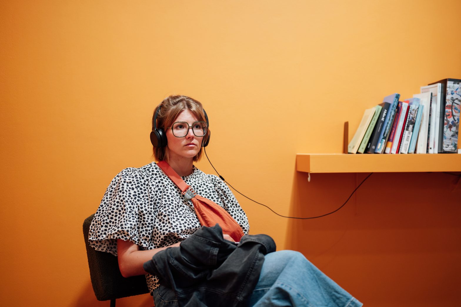 A woman sitting on a chair in front of a orange wall listening to headphones which is attached to a book shelf.