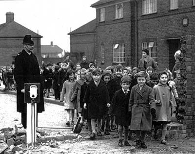 Aged black and white photo of local children standing with a policeman outside brick houses with a demolished wall.