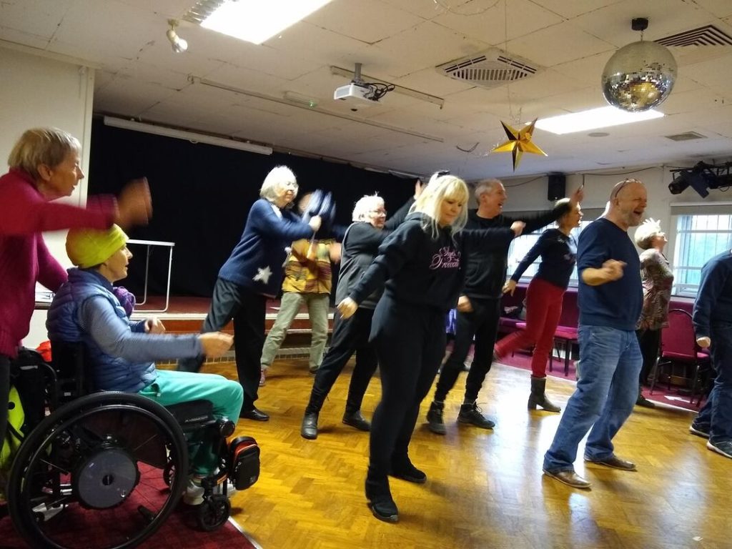A pantomime rehearsal taking place in a room with a wooden floor and a disco ball and star on the ceiling, with participants making poses with their arms and legs including one person in a wheelchair.