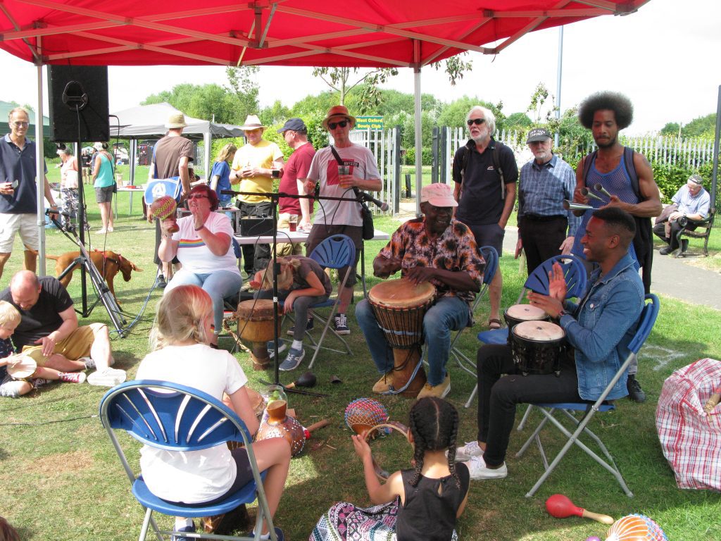 Photo of a band playing at a sunny community event run by West Oxford Community Centre. The band play with African drums and a child plays a tambourine.
