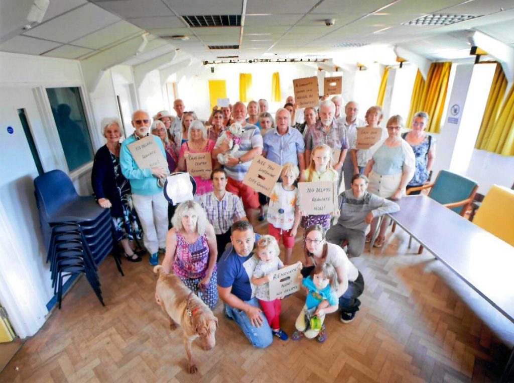Group photo from 2017 inside a room with stacked chairs and a wooden floor, with people of different ages and a dog. The people hold cardboard placards.