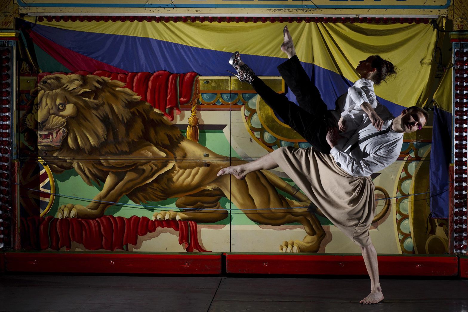 A man is lifting a woman up into the air in a dance move where both her legs are pointed to the ceiling and her arms are outstretched. Behind them a tapestry of a painting hangs from the ceiling.