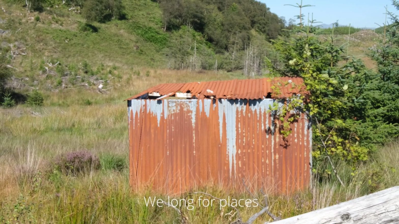 Corrugated metal hut which has gone rusty in a hilly landscape with trees and grass, with the words: We long for places