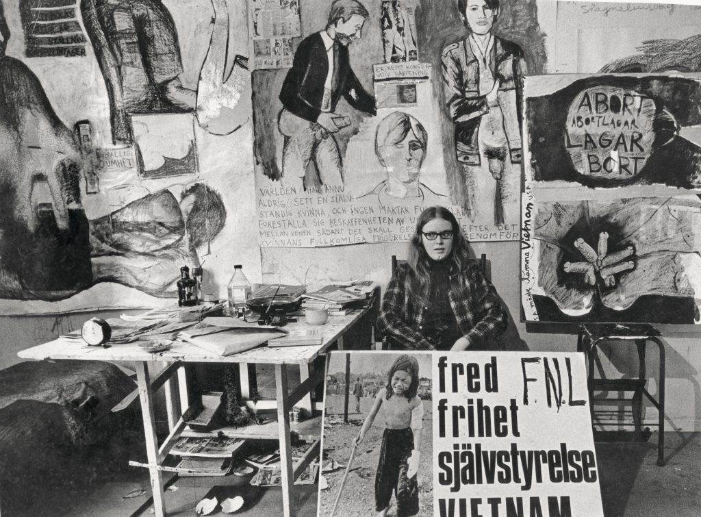 Monica Sjöö sits in her studio, surrounded by her paintings and large protest placards protesting the Vietnam War.