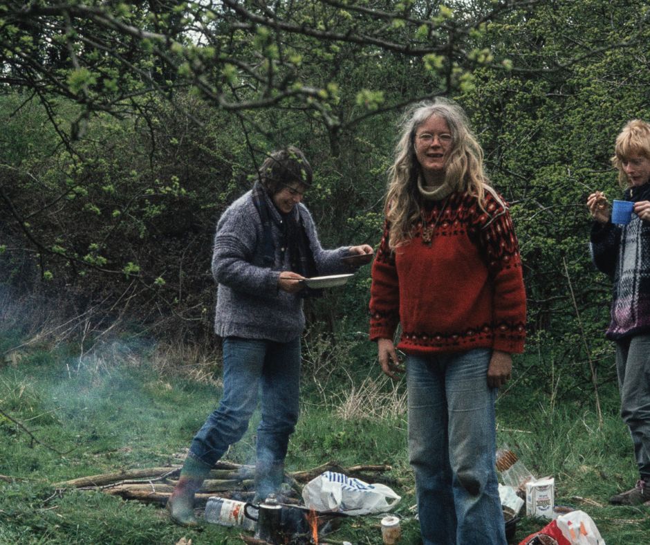A photo of three people cooking in a forest. The woman in the forefront is artist Monica Sjöö, smiling at the camera. She is wearing a red jumper and blue jeans and has white long hair.