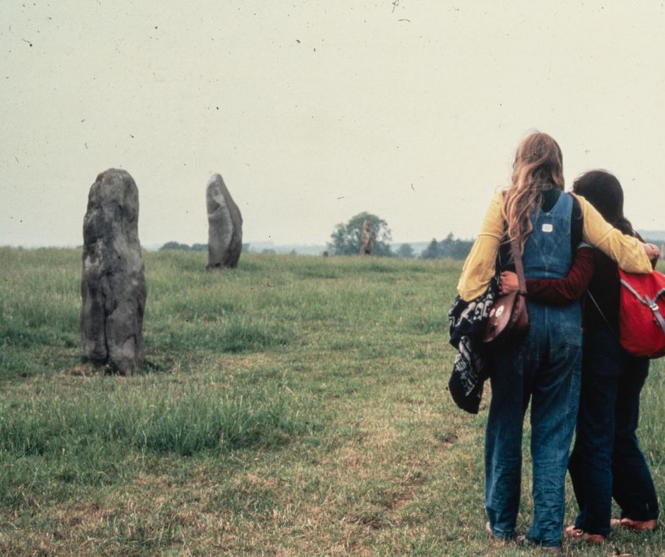 A grainy photograph of artist Monica Sjöö with her arms around another woman in front of some big stones in a field.