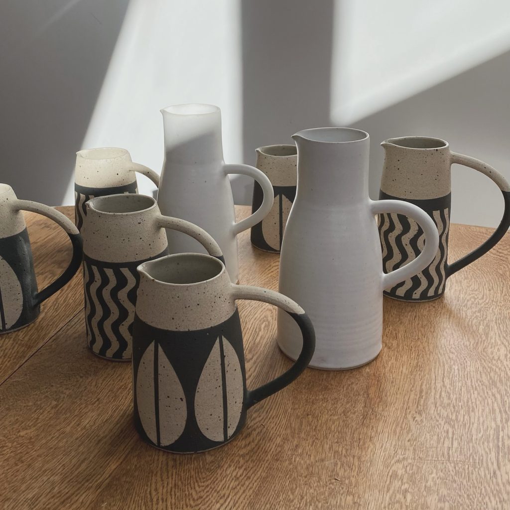 Photograph of simple, stoneware jugs in white, grey and a beige. Some have wave or leaf designs on them.