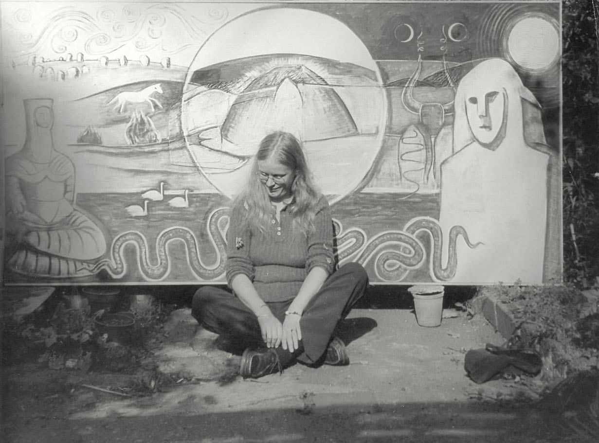 Black and white photograph of Monica Sjöö sitting cross-legged in front of one of her paintings