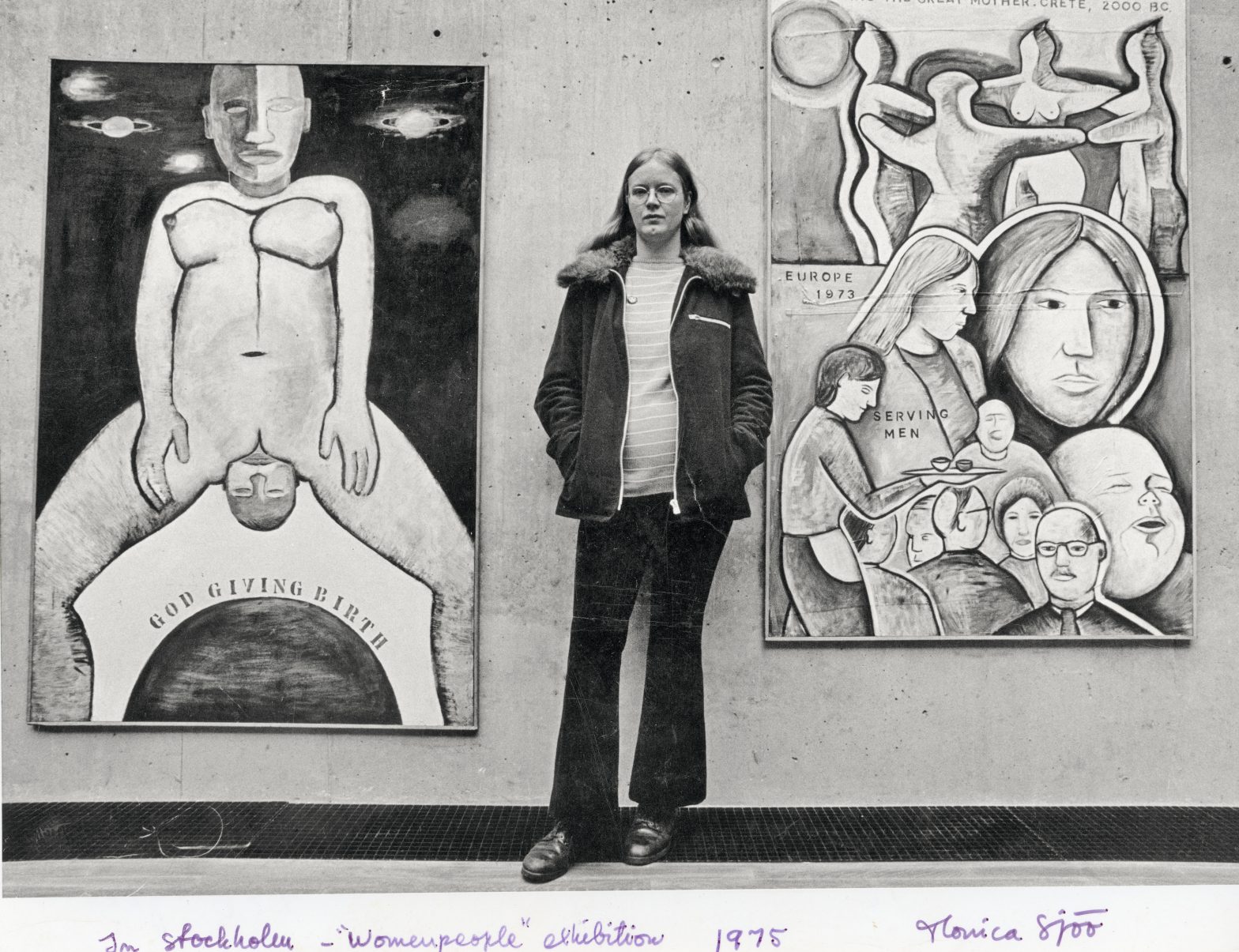 A black and white photo of artist Monica Sjöö standing in front of two of her paintings.