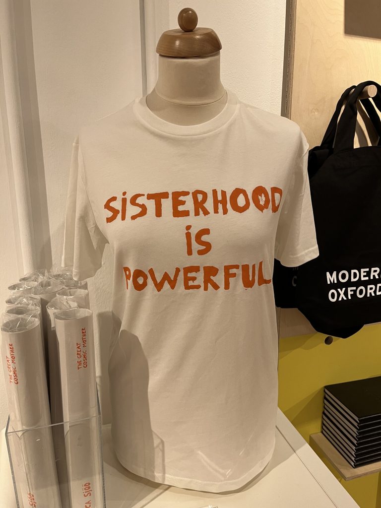 Photograph of a white cotton T-shirt with the words 'SISTERHOOD IS POWERFUL' printed on it in bright orange.