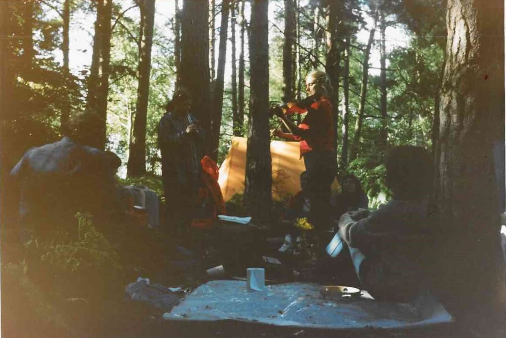 A group of women sit among trees at Green Gate, in the Greenham Common Peace Camp. Some of them play instruments, whilst others sit on the ground listening.