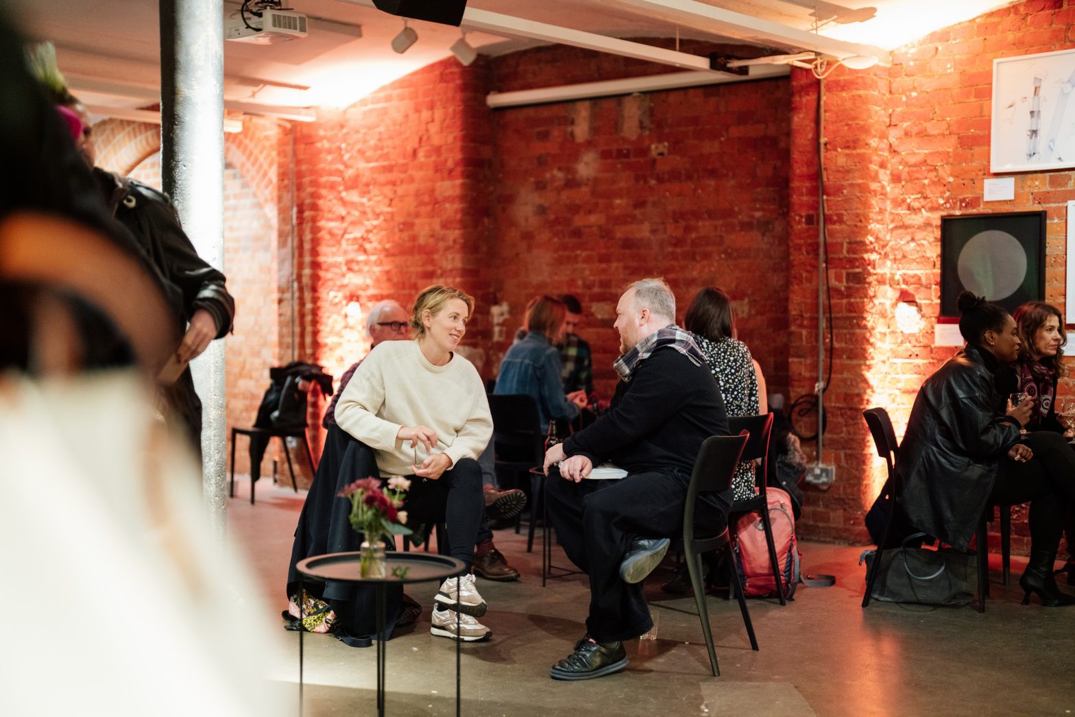 A photo of two people sitting at a black table chatting and drinking. Behind them a red brick wall is lit up by a glowing warm light and other groups of people are stood and sat around the room.