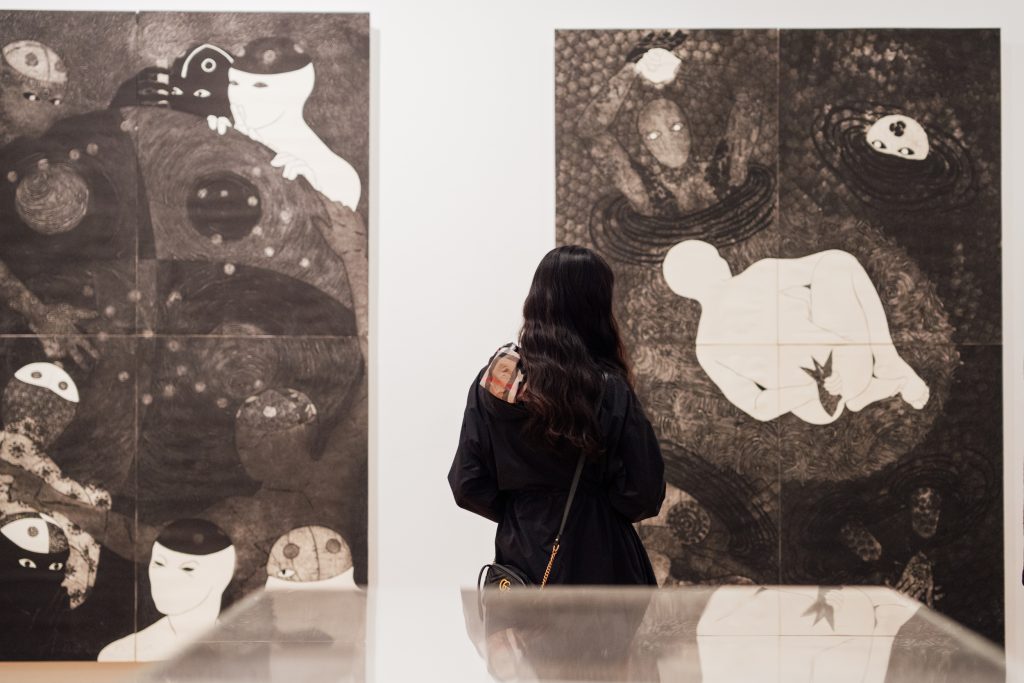 A woman with dark hair stands looking at an artwork by Belkis Ayón.