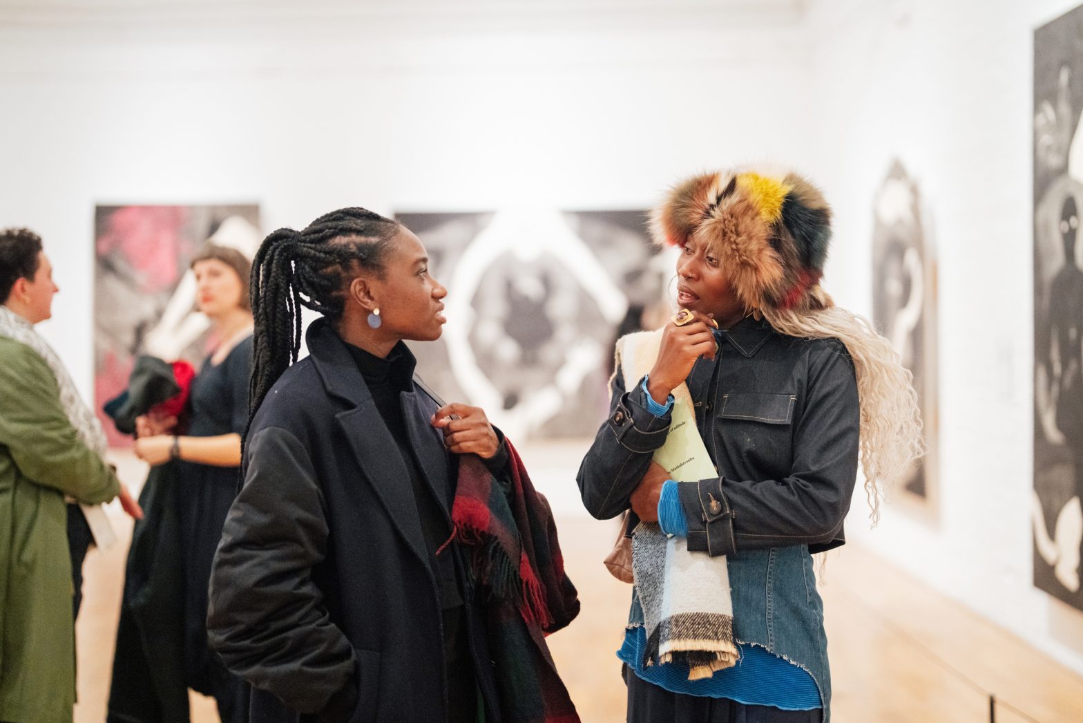 Two women are stood in a bright art gallery chatting about the paintings on the wall behind them. One is wearing a brightly coloured fluffy hat and the other woman has her hair pulled back in long braids.