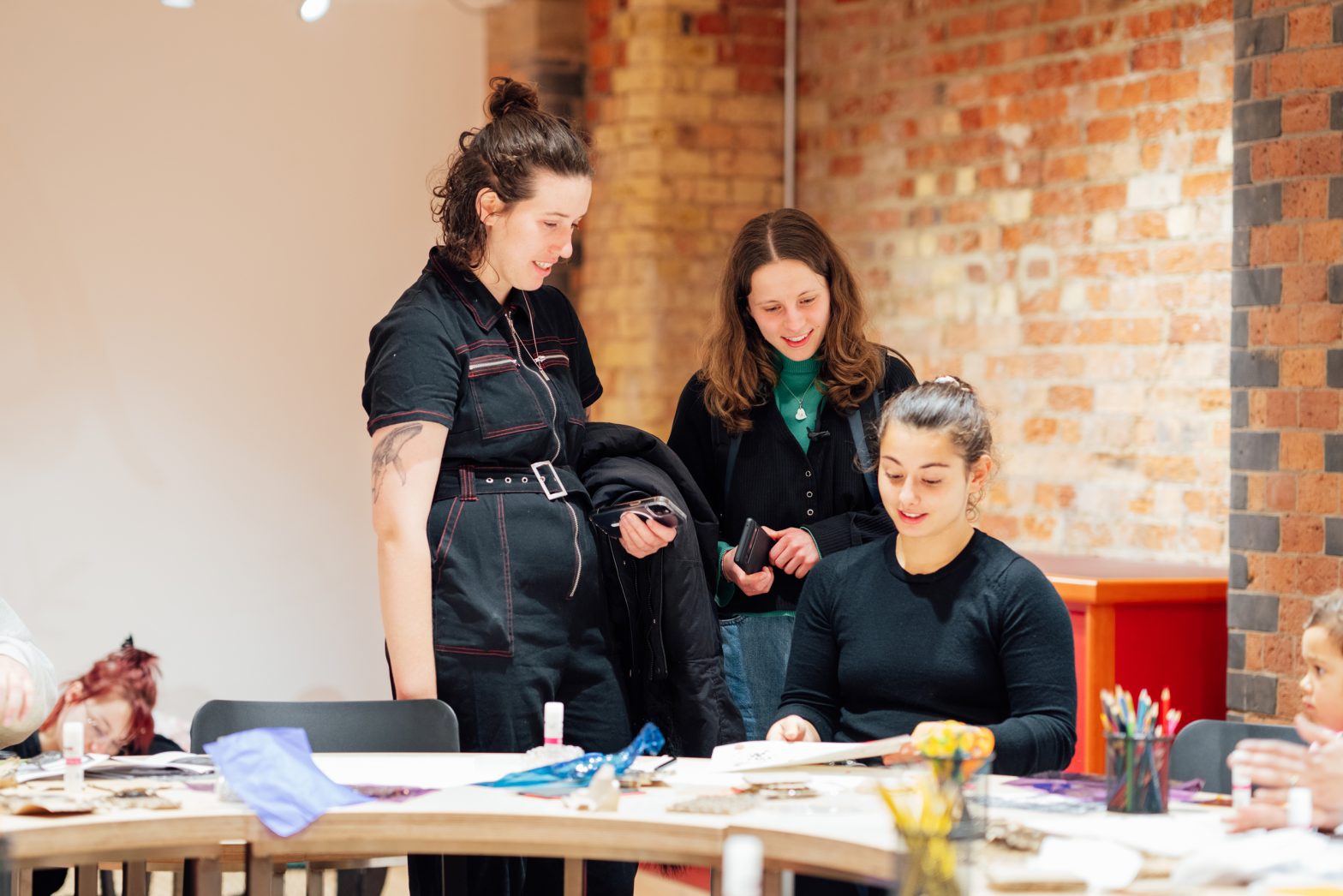 Thee women are sat around a table at a workshop laughing and chatting with each other.