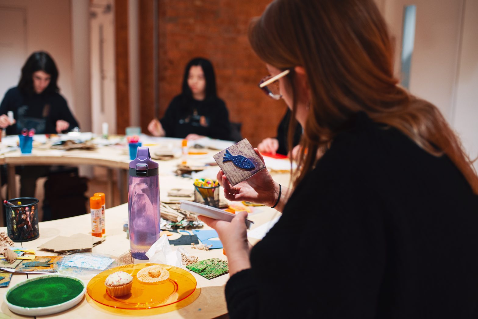 A woman is sitting around a table with other members of Modern Art Oxford's Young Creatives. They are printmaking with cardboard and ink on the table.