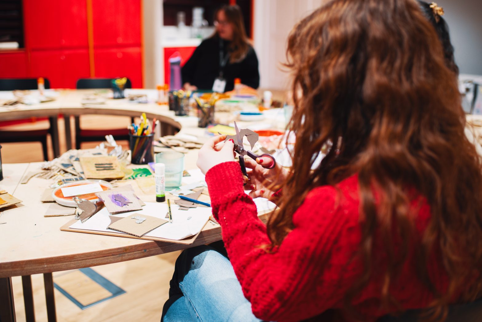 A photo of a girl cutting things out during a workshop. She is sat at a table with other young people.