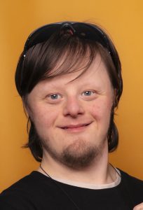 A photo of a man looking straight at the camera. He is smiling and has long brown hair and a beard. The background is yellow. 