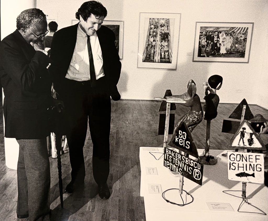 A photo of Desmond Tutu and Director David Elliot in the Art from South Africa exhibition in 1990. They are stood together, wearing suits looking at sculptures on a table. Behind them paintings are on the wall of the gallery.