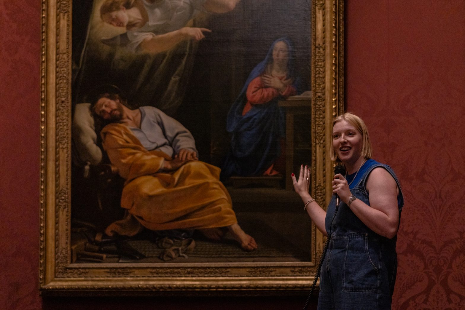 A blonde woman is standing in front of an old artwork holding a microphone and laughing
